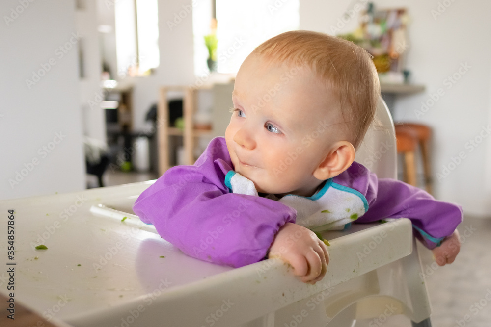 Cheerful cute baby girl sitting in tall chair spotted with green puree, looking away. Closeup shot. First solid food or child care at home concept Cheerful cute baby girl sitting in tall chair spotted with green puree, looking away. Closeup shot. First solid food or child care at home concept