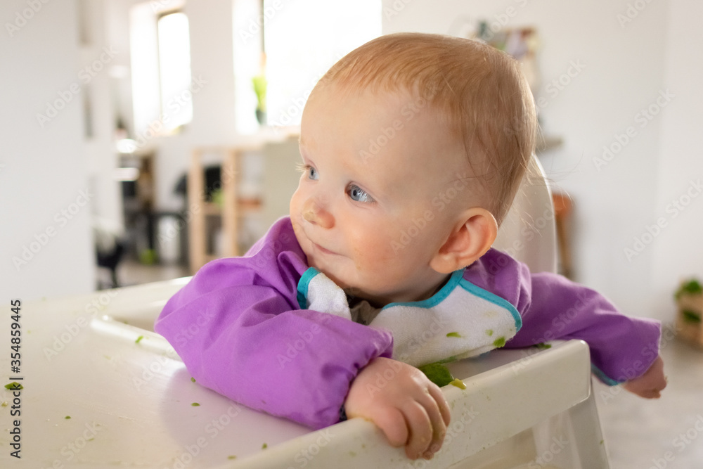 Happy funny baby girl sitting in tall chair with puree smudges, looking away. Closeup shot. First solid food or child care at home concept Happy funny baby girl sitting in tall chair with puree smudges, looking away. Closeup shot. First solid food or child care at home concept