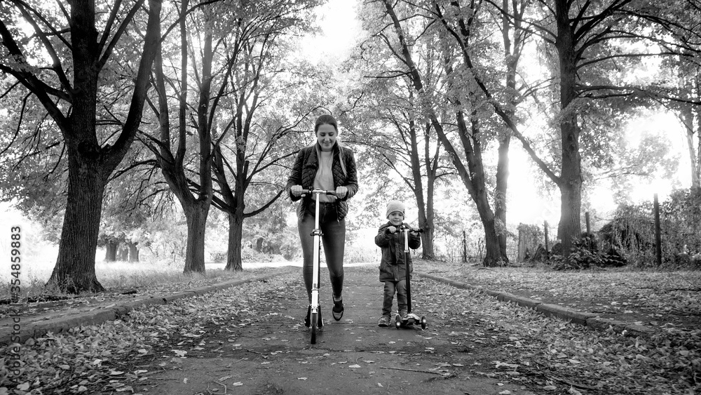 Black and white photo of young mother with little son riding kick scooter on long alley at autumn park Black and white photo of young mother with little son riding kick scooter on long alley at autumn park