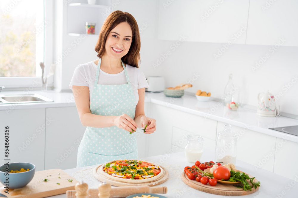 Portrait of positive charming girl housewife cook tasty pizza seasoning oregano wear dotted apron in house indoors