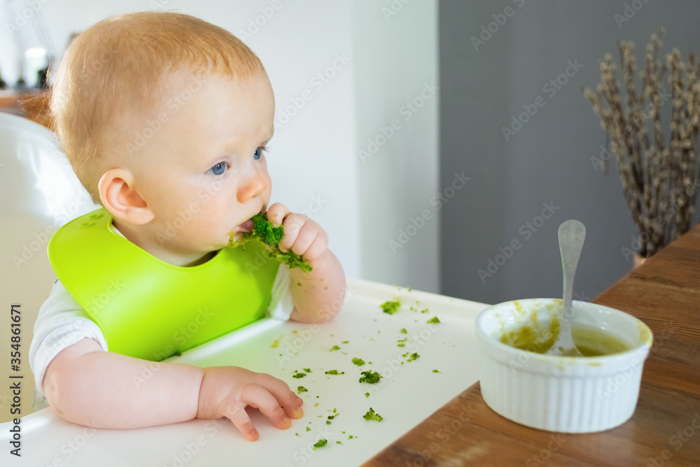 Pensive baby eating broccoli vegs and soup. Little child wearing plastic bib, sitting in highchair. First solid food or child care at home concept Pensive baby eating broccoli vegs and soup. Little child wearing plastic bib, sitting in highchair. First solid food or child care at home concept