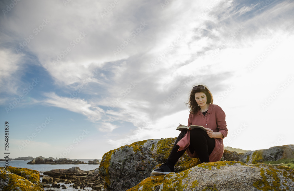 Black-haired woman reading book on the rocks by the sea