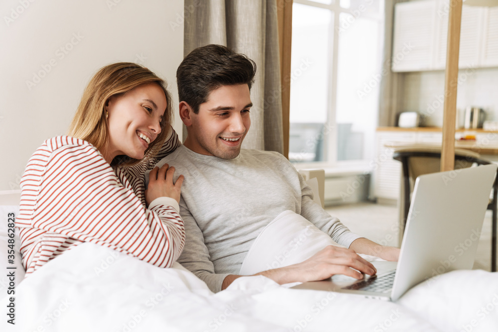 Portrait of joyful couple smiling and using laptop while lying in bed Portrait of joyful couple smiling and using laptop while lying in bed