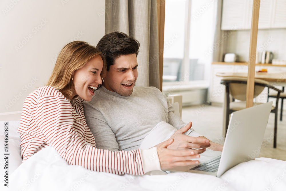 Portrait of excited couple smiling and using laptop while lying in bed Portrait of excited couple smiling and using laptop while lying in bed