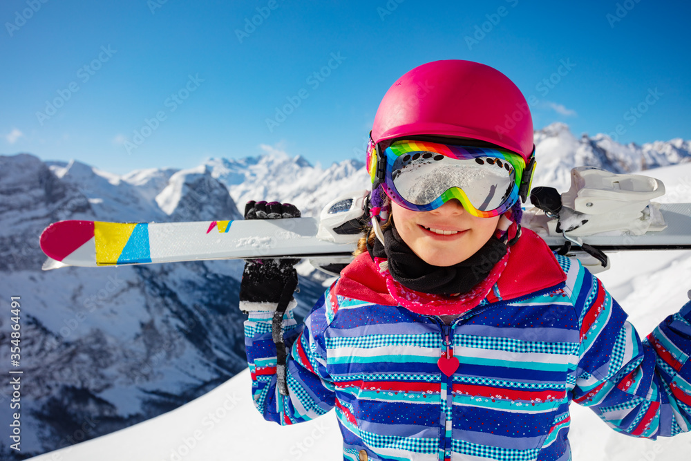 Close-up face portrait of a girl in colorful outfit, pink helmet and color glasses hold ski on her shoulders view over mountain summit on background