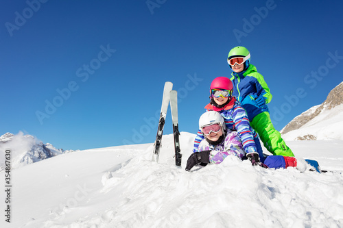 Pile of kids one on top of another lay in the snow over blue sky in the mountains with ski on background