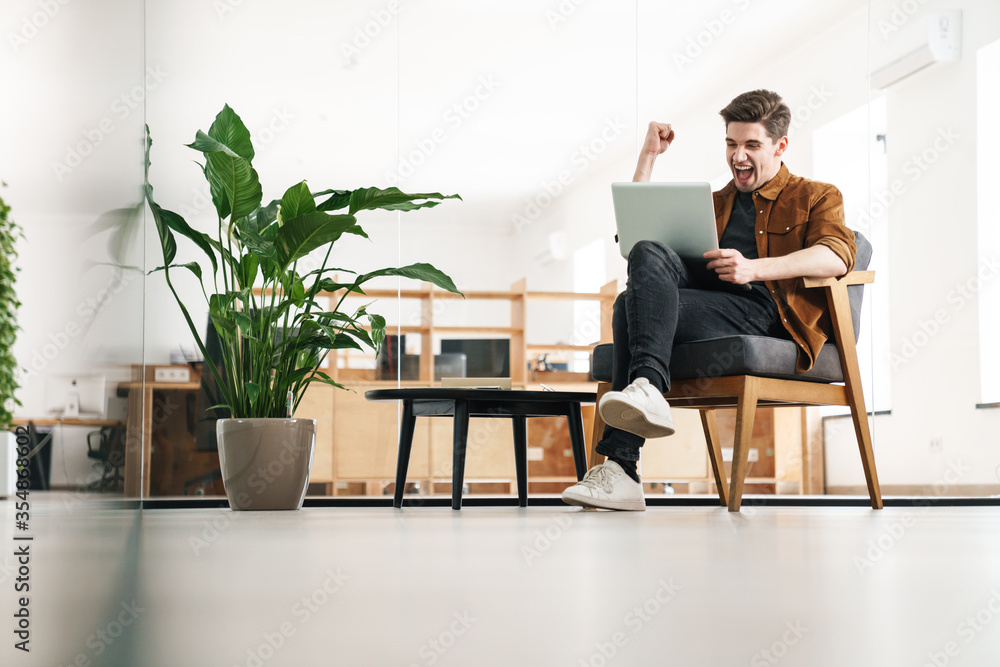 Image of delighted man making winner gesture while working with laptop Image of delighted man making winner gesture while working with laptop