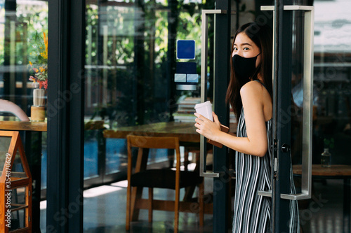 Young beautiful Asian woman wearing a surgical face mask while leaving a coffee cafe shop. Attractive female customer buying coffee and holding a disposable coffee cup. Covid-19 prevention.