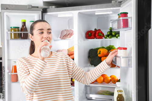 Young woman drinking water near open refrigerator indoors