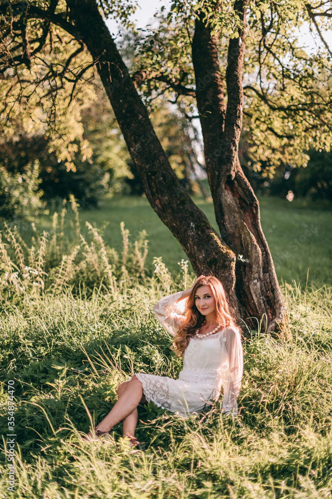 Portrait of a beautiful young blond woman in a white dress in a boho style walks in the park in summer at sunset.