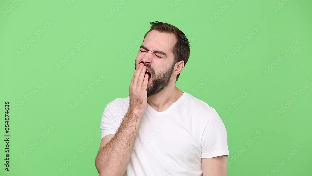 Tired bearded young guy 20s in white t-shirt isolated on green chroma key background studio. People sincere emotions lifestyle concept. Looking at camera yawning want to sleep cover mouth with hand Tired bearded young guy 20s in white t-shirt isolated on green chroma key background studio. People sincere emotions lifestyle concept. Looking at camera yawning want to sleep cover mouth with hand