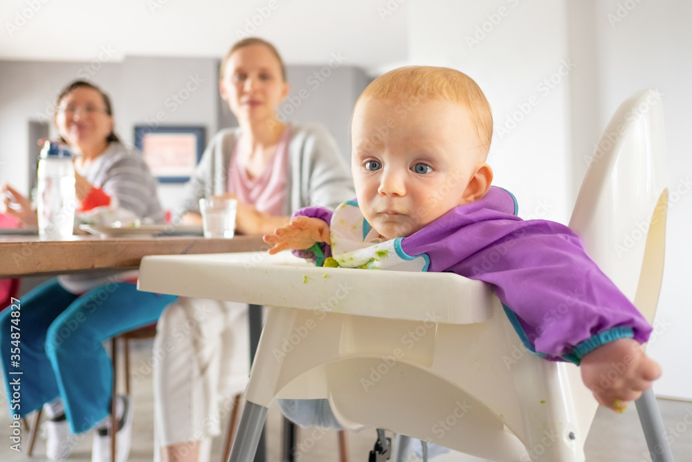 Sweet red haired baby having dinner with family. Little child sitting in high chair with puree smudges on tray. Feeding or child care at home concept Sweet red haired baby having dinner with family. Little child sitting in high chair with puree smudges on tray. Feeding or child care at home concept