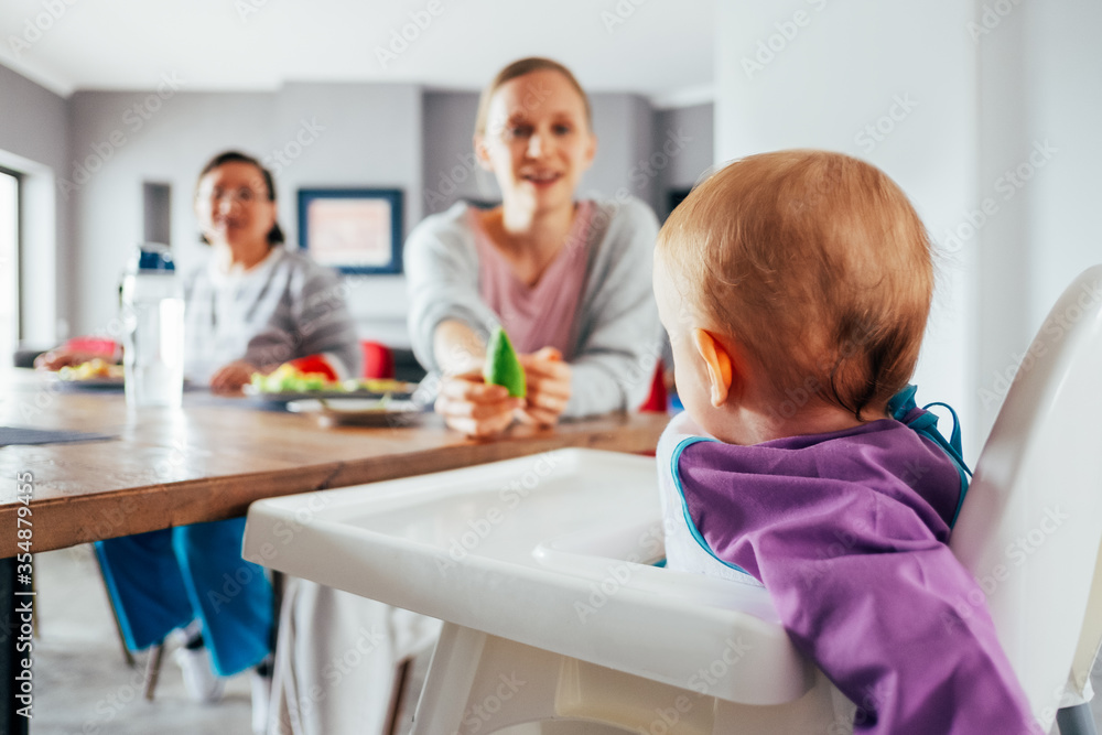 Young mom feeding her child with solid food in dining room. Cute baby girl sitting on highchair and having breakfast. Childcare and led weaning concept