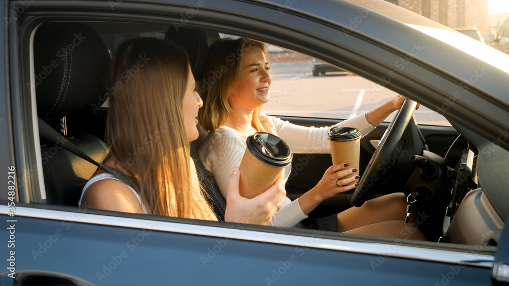 Naklejka premium Portrait of two smiling girls driving in car and drinking coffee from paper cups at early morning