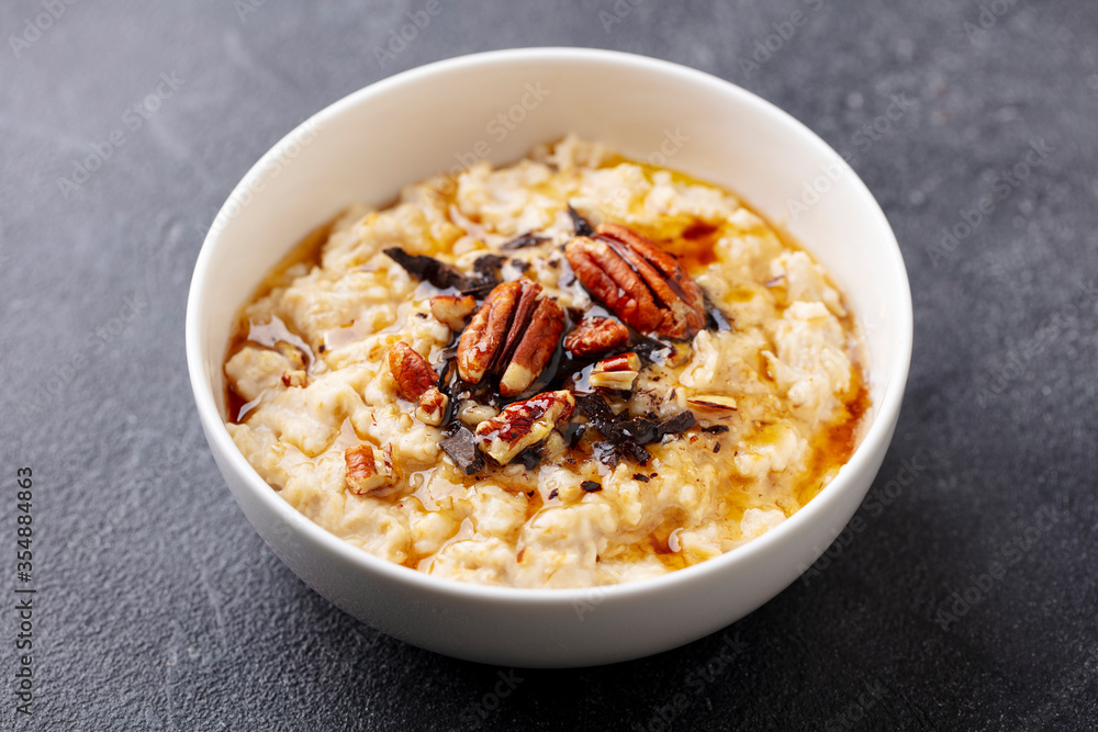 Oatmeal porridge with pecan nuts, maple syrup and chocolate in white bowl. Grey background. Close up. Oatmeal porridge with pecan nuts, maple syrup and chocolate in white bowl. Grey background. Close up.