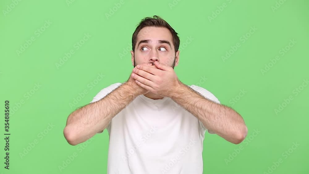 Bearded young guy 20s in white t-shirt isolated on green chroma key background studio. People emotions lifestyle concept. Looking at camera covering mouth with hands arms do not tell them information Bearded young guy 20s in white t-shirt isolated on green chroma key background studio. People emotions lifestyle concept. Looking at camera covering mouth with hands arms do not tell them information