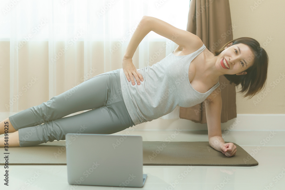Woman is doing side plank exercises, looking at the laptop in the living room on floor mat at home. Woman is doing side plank exercises, looking at the laptop in the living room on floor mat at home.