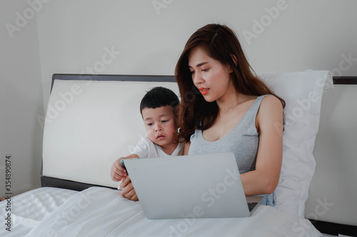 The mother works with her son in the bedroom at home. work at home, concept with a women sitting on the bed with laptop and holding a coffee mug.