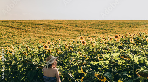 A young blonde woman with short hair in a hat and blue dress stands in a field near yellow ripe sunflowers with seeds in summer at sunset.