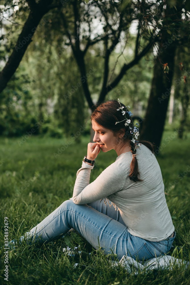 Portrait of young white beautiful woman with small fresh flowers in hair and pigtails from back sits on grass in nature in summer in garden at sunset. Nature, beautiful rays of light, park. Portrait of young white beautiful woman with small fresh flowers in hair and pigtails from back sits on grass in nature in summer in garden at sunset. Nature, beautiful rays of light, park.