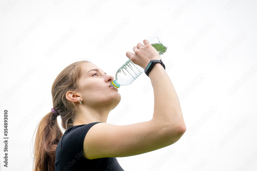 Beautiful girl drinks water from a bottle after a workout in the park. On the grassland the reflection of green grass