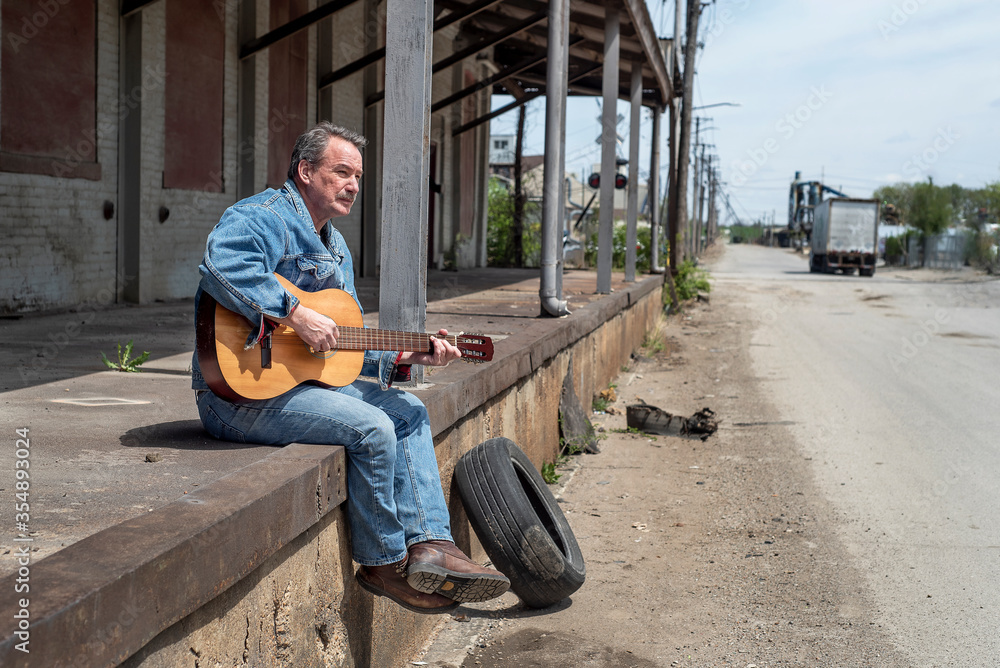 Man playing acoustic guitar wearing jeans outside in the city sitting on old loading dock Man playing acoustic guitar wearing jeans outside in the city sitting on old loading dock