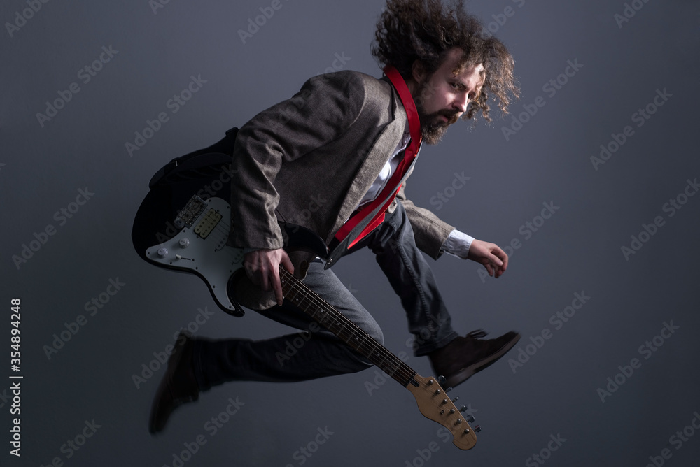 Obraz premium defocused studio portrait of a young bearded, curly-haired guy with long hair, in a jacket, jumping with a guitar, looking at the camera. On a gray background. Dynamic male dramatic portrait
