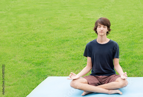 A young man with long hair does yoga outdoors. A teenager sits in a lotus position on the green grass. Copy space. Place for text