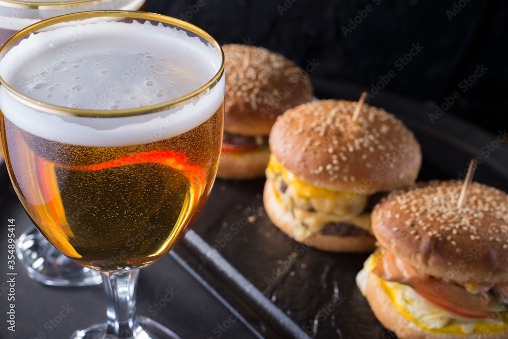 close-up, beer glasses with craft dark and light beer with foam, against a background of burgers, with a variety of ingredients, including shyomga, beef meat patty, cheese burger. In a rustic style close-up, beer glasses with craft dark and light beer with foam, against a background of burgers, with a variety of ingredients, including shyomga, beef meat patty, cheese burger. In a rustic style