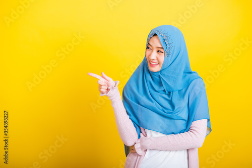 Asian Muslim Arab, Portrait of happy beautiful young woman Islam religious wear veil hijab funny smile she positive expression pointing finger side sideways to space isolated yellow background © sorapop