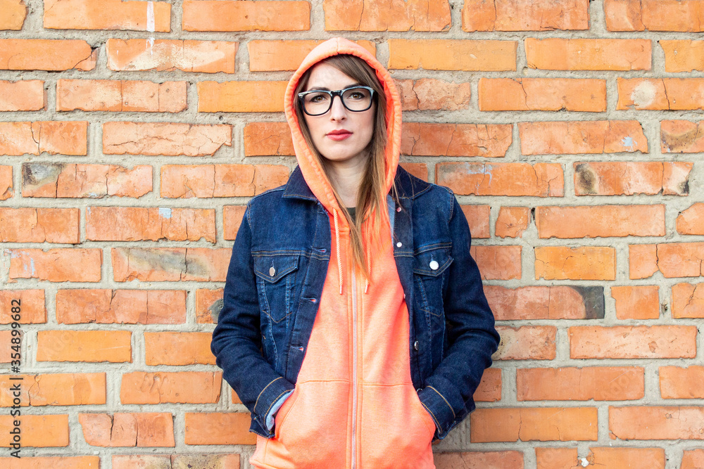 Serious young girl wearing glasses, arms crossed, wearing a hoodie, wearing a hood over his head and a denim jacket on a red brick wall