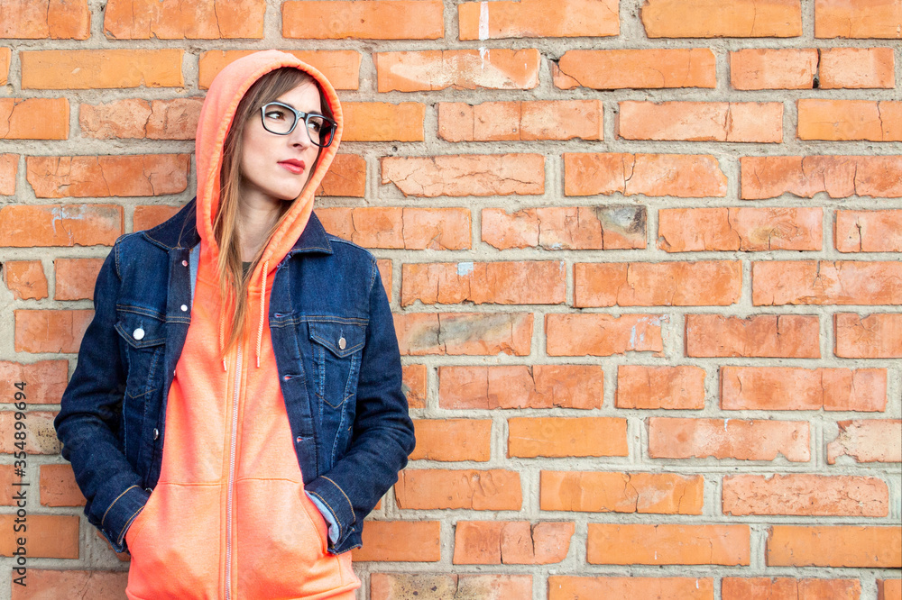 Serious young woman with glasses, dressed in a sweatshirt and denim jacket, hands holding a sweatshirt in her pocket, looking to the side against a red brick wall