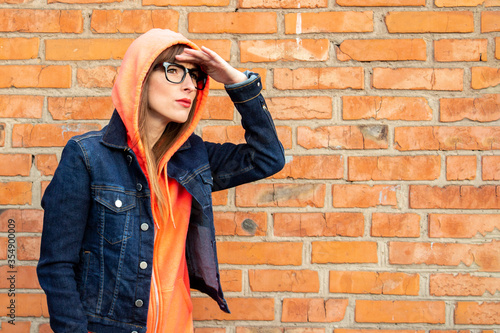 Young girl with glasses, put his hand looking into the distance, wearing a hoodie and denim jacket on a red brick wall