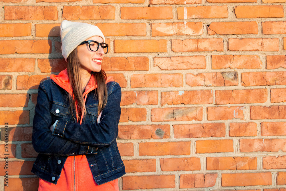Young woman smiling with his arms crossed looking to the side wearing glasses on the background of a brick wall, wearing a gray cap, hoodie and denim jacket