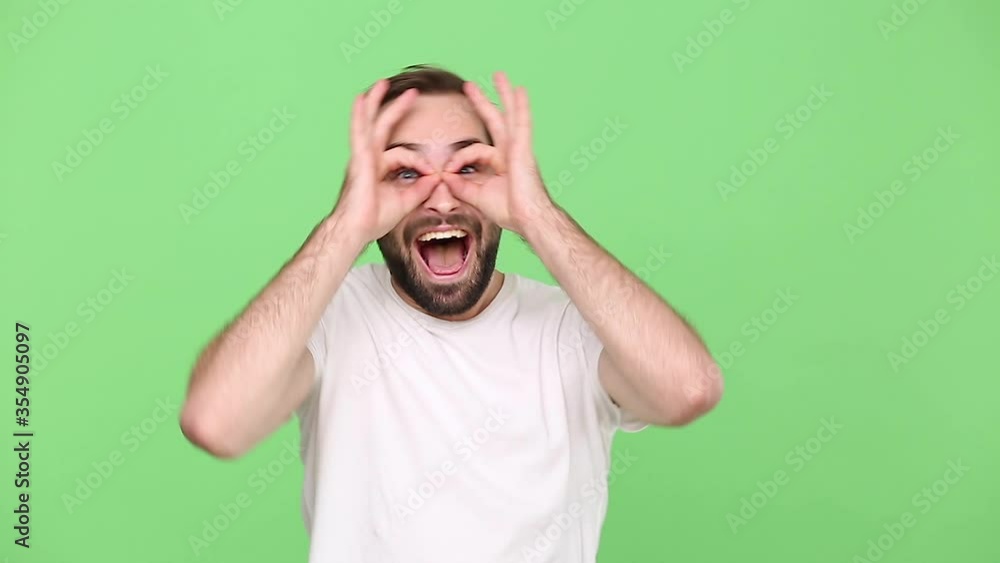 Bearded young guy 20s in white t-shirt isolated on green chroma key background studio. People lifestyle concept. Looking camera fooling around holding hands near eyes imitating glasses or binoculars Bearded young guy 20s in white t-shirt isolated on green chroma key background studio. People lifestyle concept. Looking camera fooling around holding hands near eyes imitating glasses or binoculars
