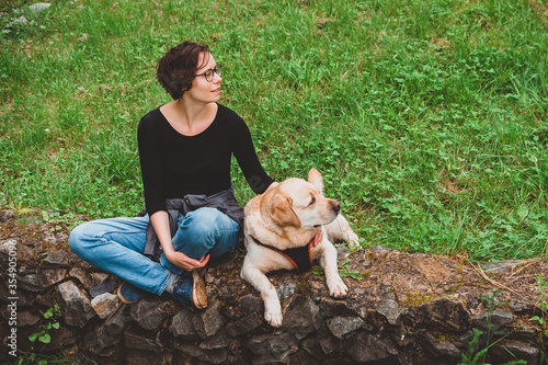 The girl is sitting in an embrace with a Labrador Retriever on the grass. Rest with your dog on a walk in the Park