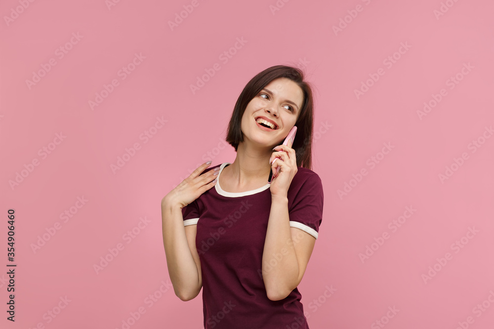 Young people working with mobile devices. Portrait of cute happy brunette woman wearing t-shirt talking on mobile phone and smiling isolated over pink background. Copy space. Young people working with mobile devices. Portrait of cute happy brunette woman wearing t-shirt talking on mobile phone and smiling isolated over pink background. Copy space.