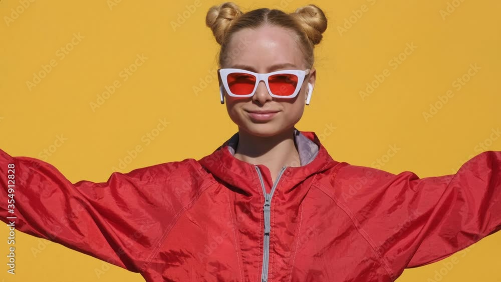 Portrait of happy teen girl blonde in airpods in stylish sunglasses, red hoodie smiles, dances, rhythmically shaking his head camera on yellow background slow motion summer. Emotions. Girl dancing Portrait of happy teen girl blonde in airpods in stylish sunglasses, red hoodie smiles, dances, rhythmically shaking his head camera on yellow background slow motion summer. Emotions. Girl dancing