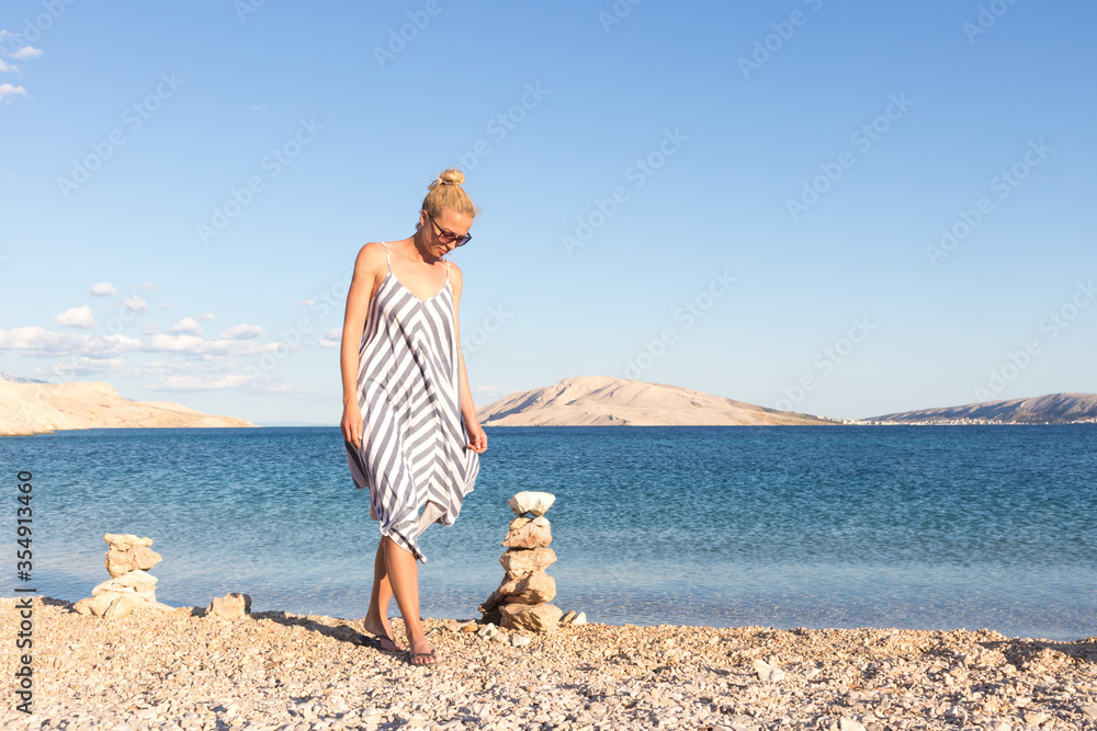 Happy carefree woman wearing beautiful striped summer dress enjoying late afternoon walk on white pabbled beach on Pag island, Croatia. Happy carefree woman wearing beautiful striped summer dress enjoying late afternoon walk on white pabbled beach on Pag island, Croatia.
