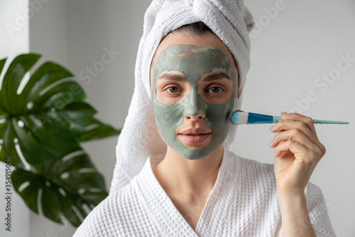 Tableau sur toile Young woman wearing towel on head and bathrobe putting clay mask on face with brush