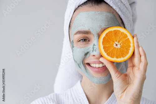 Smiling happy woman with grey clay facial mask applied on face holding orange citrus fruit slice.