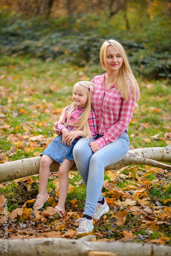 Mom and daughter in pink shirts and blue jeans are sitting on a tree in the autumn park.