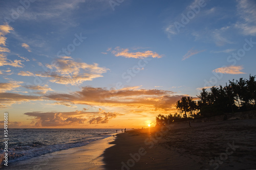 Silhouettes of walking people in beautiful sunset on the beach with palm trees. Magical golden sunset sky with clouds