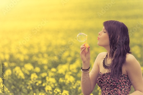 Beautiful woman blowing a single soap bubble in summer nature. The girl has fun in a yellow meadow and is enjoying her youth