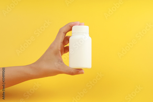Female hand holding a white plastic medicine bottle on yellow background