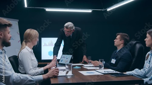 Corporate Meeting Room: Senior Confident Businessman Decisively Leans on a Conferense Table, Delivers Report and Holds Discussion with the Team of Coworkers. Elderly Executive Director Showing photo