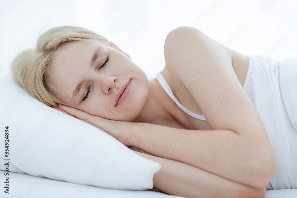 Beautiful young caucasian woman peacefully sleeping sideways on a soft comfortable white pillow and bed at home in the morning.
