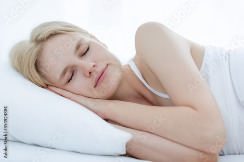 Beautiful young caucasian woman peacefully sleeping sideways on a soft comfortable white pillow and bed at home in the morning.
