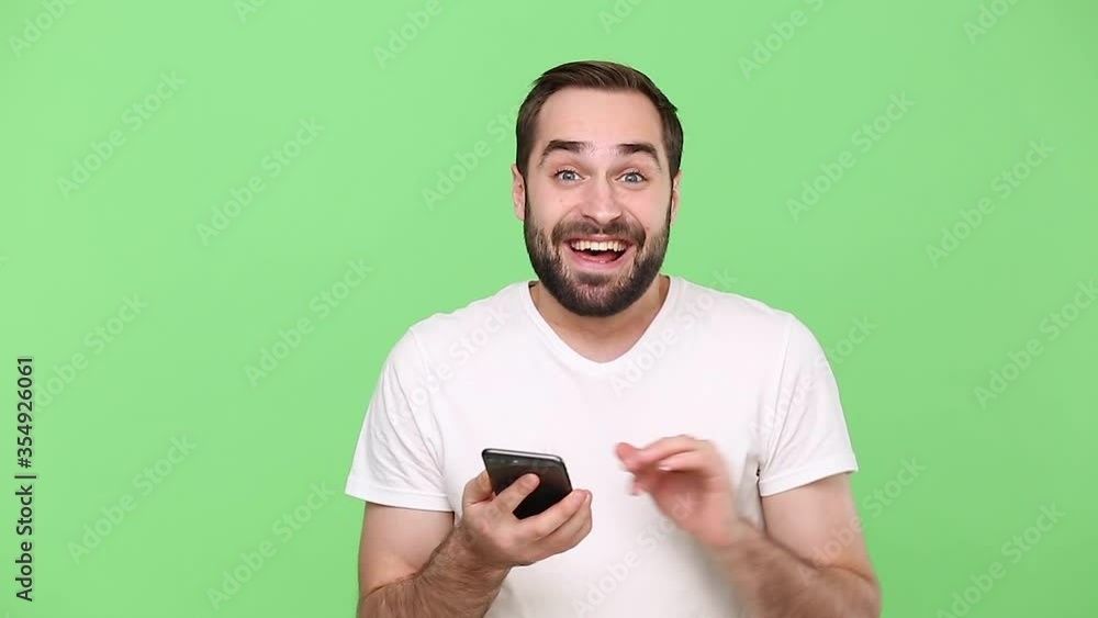 Cute young guy 20s in white t-shirt hold in hand using mobile cell phone send sms girlfriend waving hand as greet meet someone isolated on green chroma key background studio. People lifestyle concept Cute young guy 20s in white t-shirt hold in hand using mobile cell phone send sms girlfriend waving hand as greet meet someone isolated on green chroma key background studio. People lifestyle concept