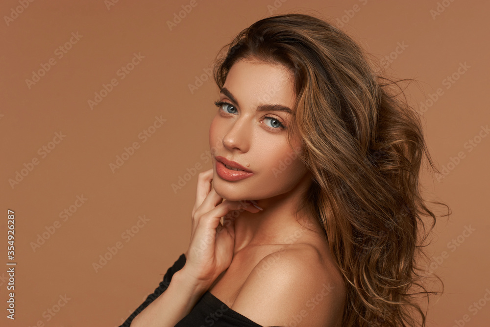Naklejka premium Girl in black dress with wavy hair and make-up posing on a beige background. Fashion style closeup studio portrait. Elegant fashionable woman
